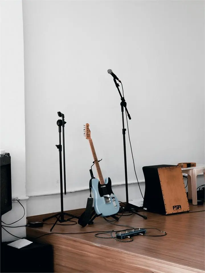 A bare practice corner with a guitar, microphone stands and a cajon on a plain floor